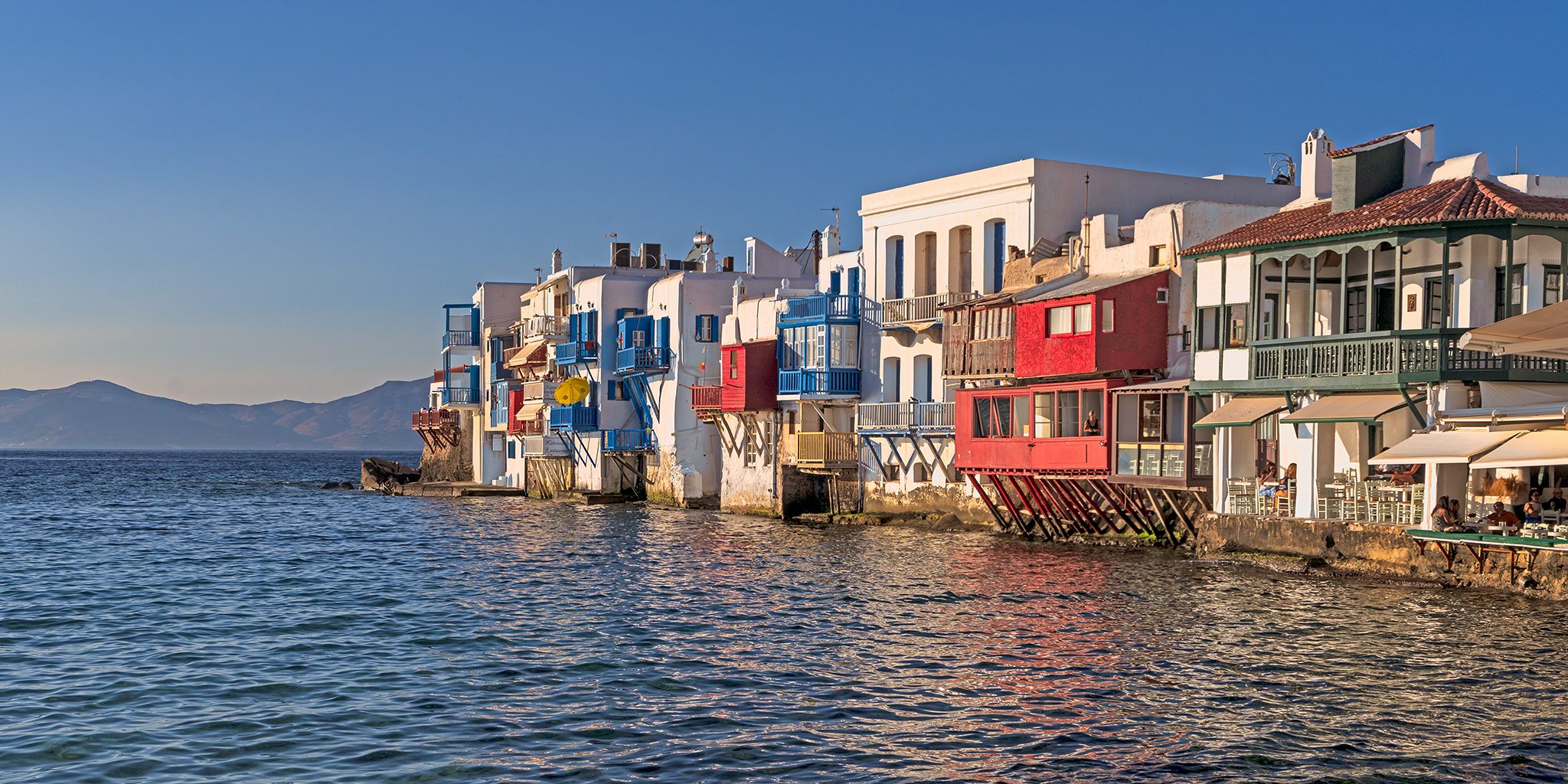 A row of white, red and blue waterfront houses with wooden balconies built right at the edge of calm sea, hills on the far side of the water.