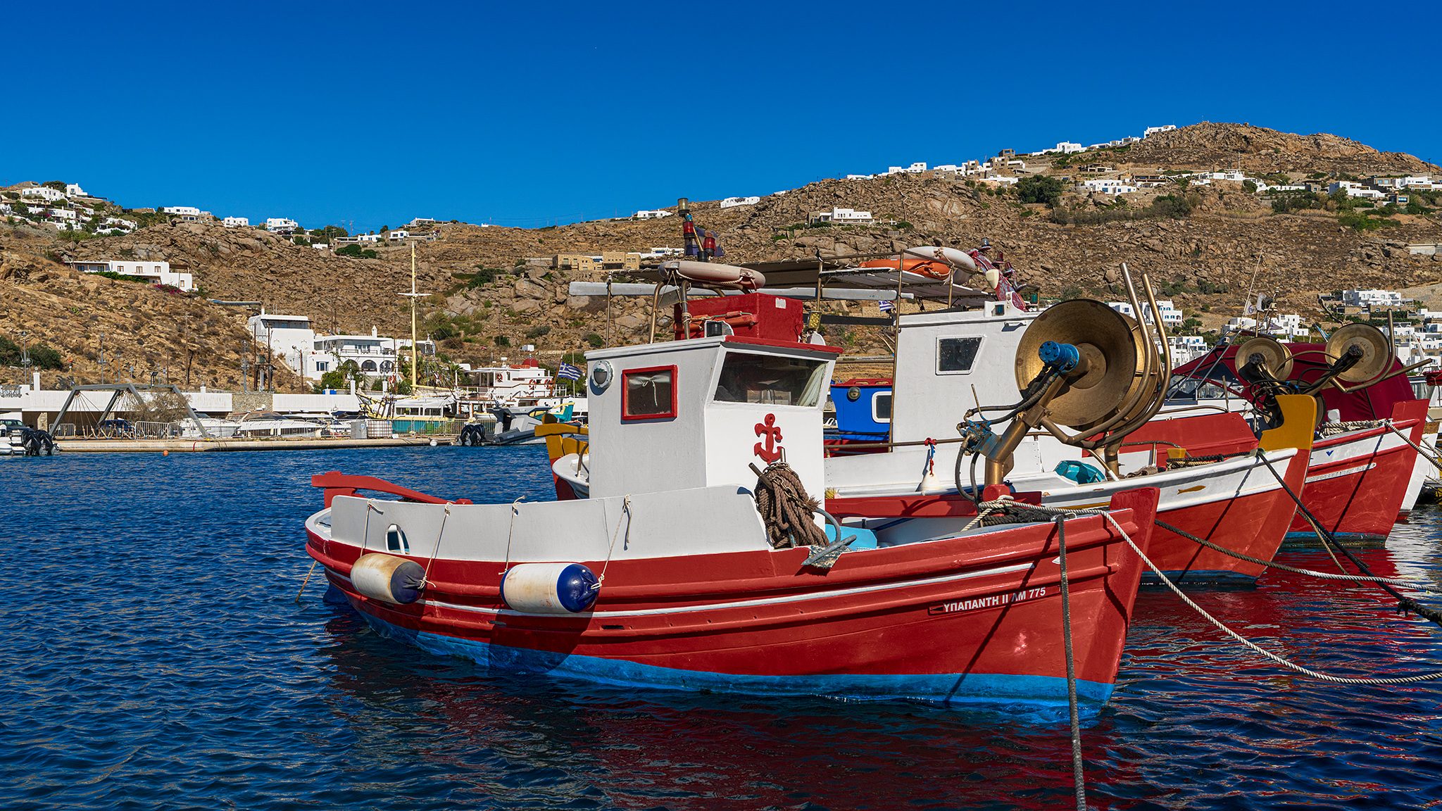 Red and white wooden fishing boats moored together in deep blue harbor water, with white houses scattered over a dry hillside behind.